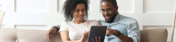 Photo of a woman and man sitting together on a tan sofa looking down at a mobile tablet