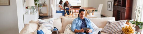 Photo of a man sitting on a tan couch with his family sitting at a brown wooden table in the back of the room