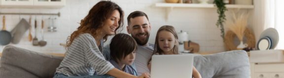 Photo of a man, his wife, and two children sitting together on a grey couch looking down at a laptop screen